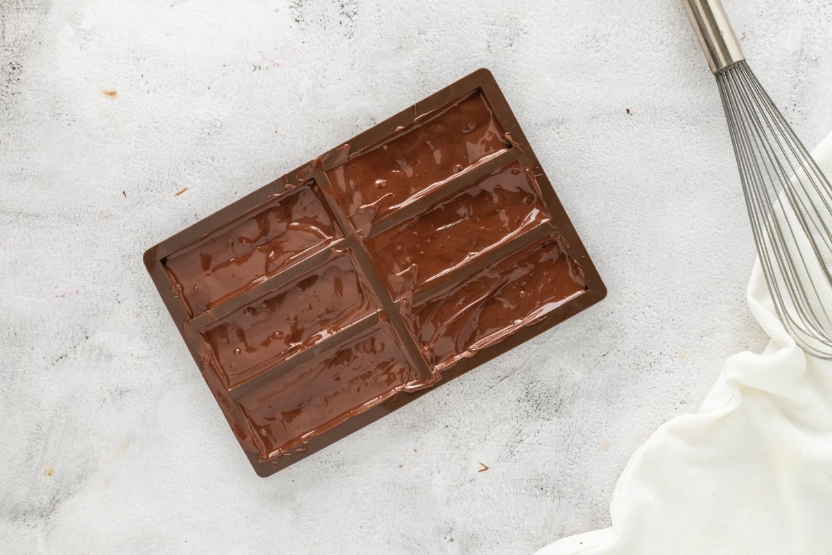 Chocolate bar mold filled with melted chocolate, whisk and cloth beside it.