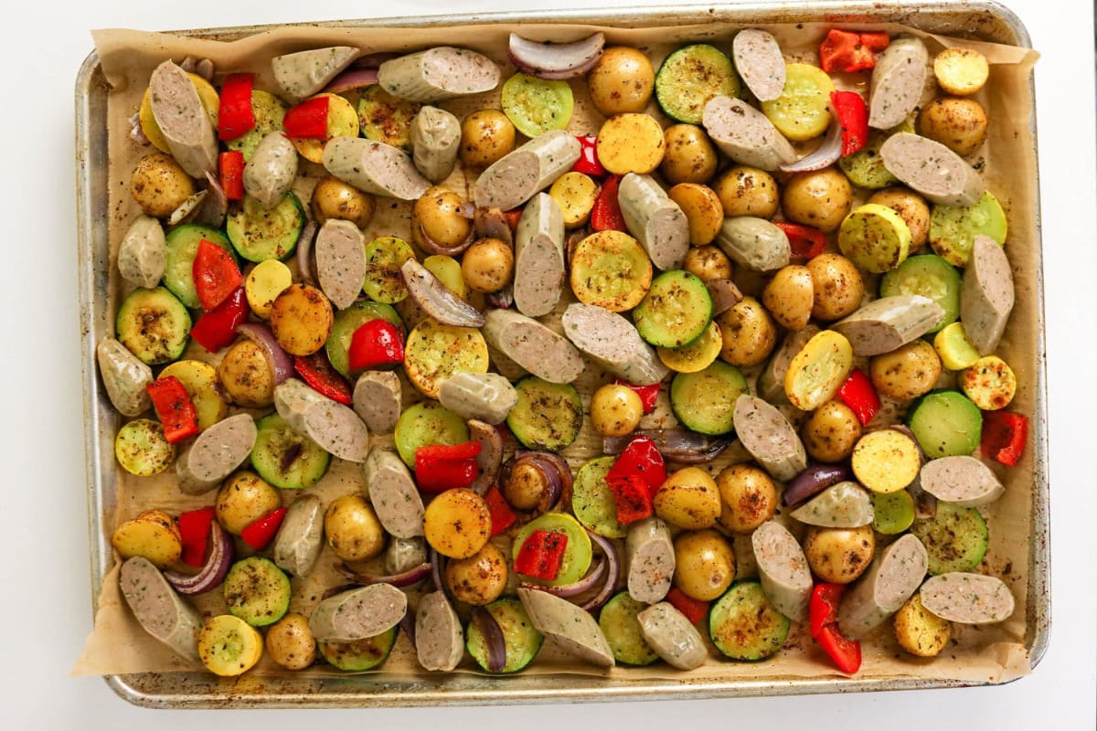 Sheet pan with sliced sausage, potatoes, zucchini, red pepper, and seasonings.