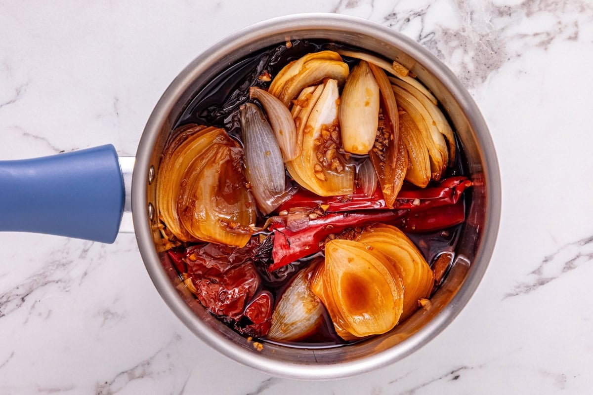 Sliced onions, dried chilies, and sauce simmering in a saucepan on marble.