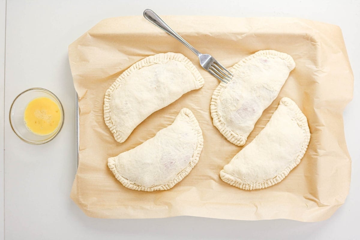 Four unbaked hand pies on parchment with a fork and egg wash in a small bowl.