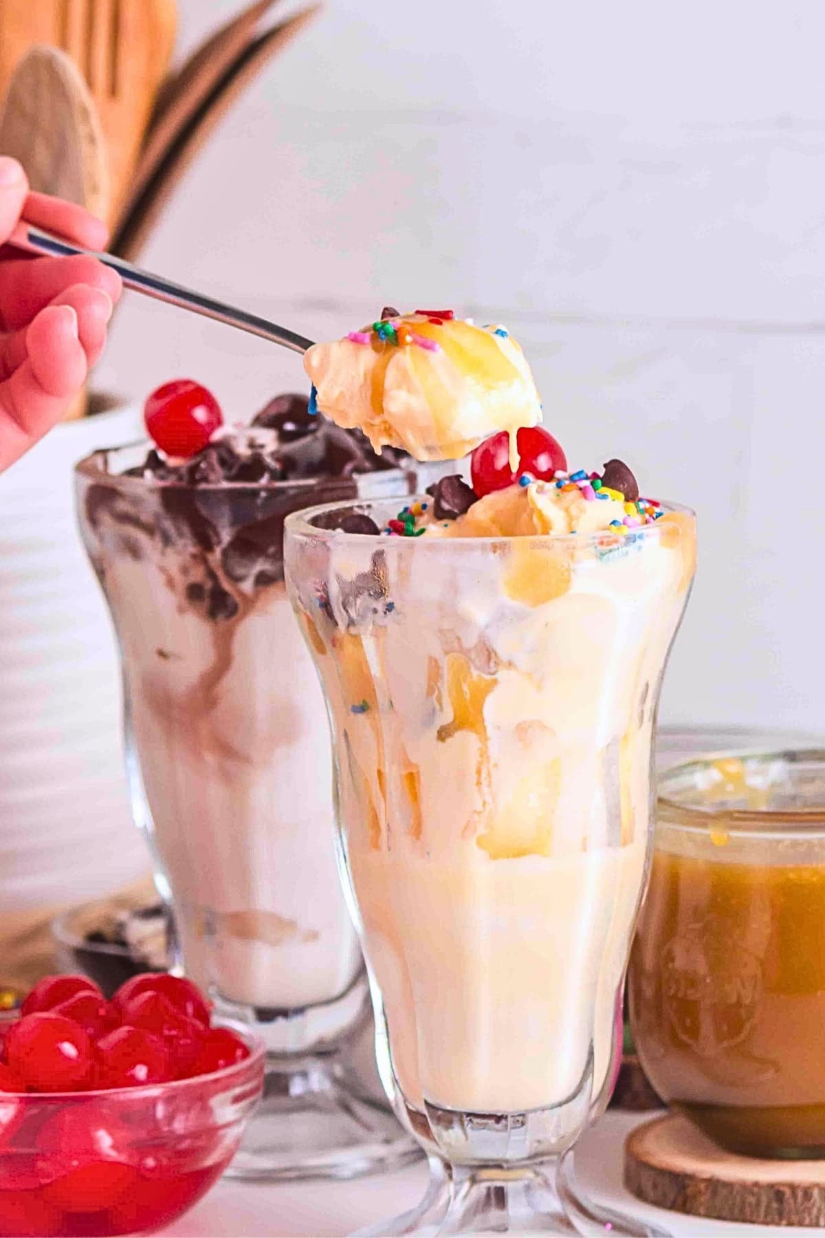 A hand holds a spoonful of ice cream sundae with cherries and sprinkles.