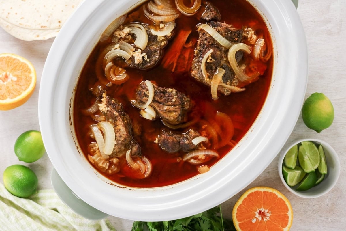 Slow cooker with braised meat, onions, broth, limes, and oranges.