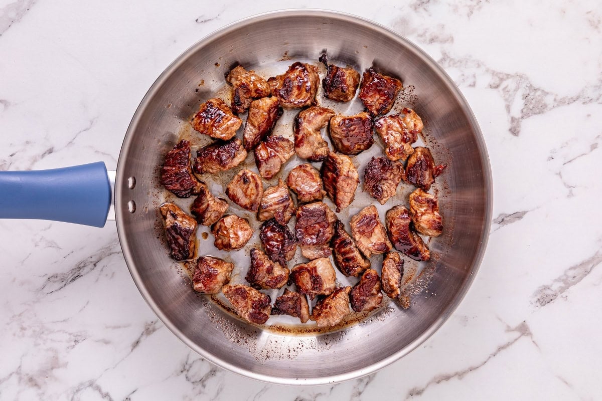 Saut&eacute;ed beef cubes cooking in a stainless steel skillet on a marble countertop.