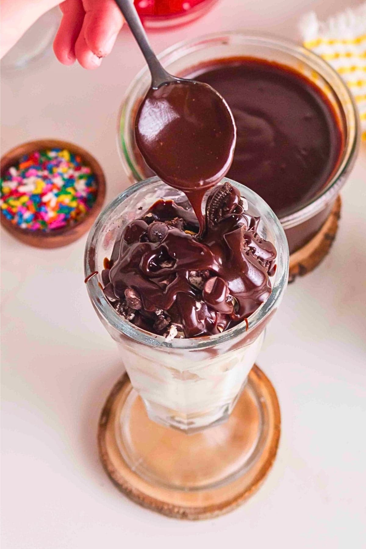 A spoon drizzles chocolate sauce over a glass of ice cream with cookies.