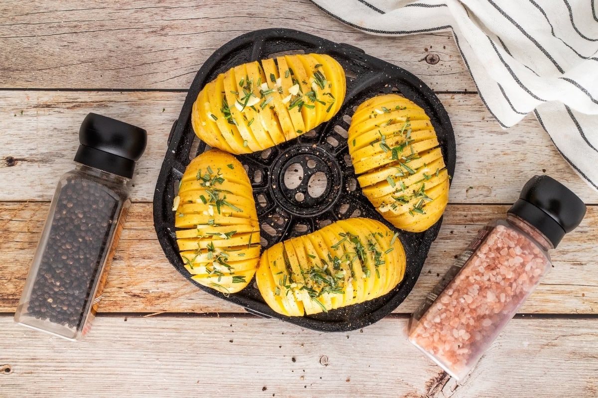Four sliced baked potatoes with herbs, pepper, and pink salt on a wooden table.