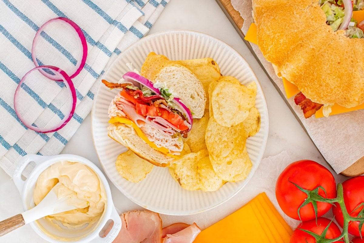 A sandwich with chips on a plate, surrounded by tomato, cheese, and onions.