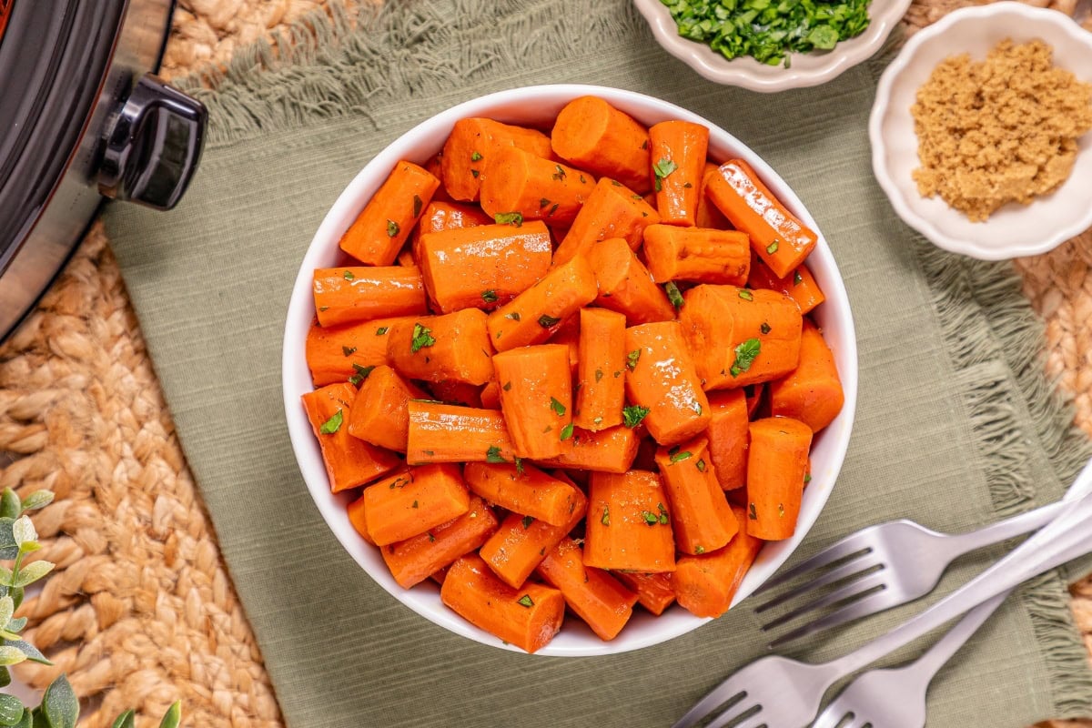 Bowl of cooked carrots garnished with herbs, next to forks and small dishes.