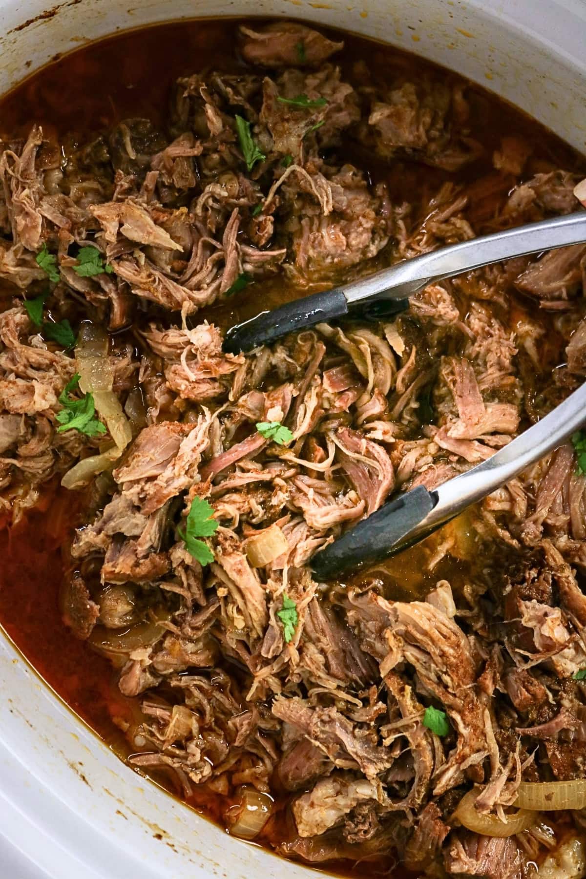 Tongs lifting shredded beef in a slow cooker with sauce and herbs.