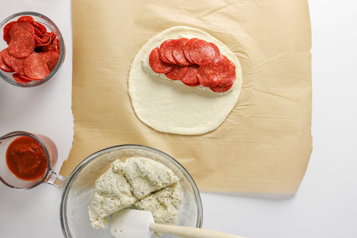Pizza dough topped with cheese and pepperoni on parchment, sauce on the side.
