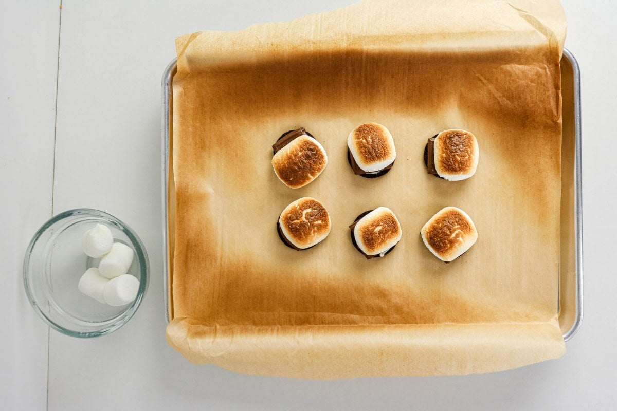 Six toasted smores on parchment with a glass cup of marshmallows nearby.