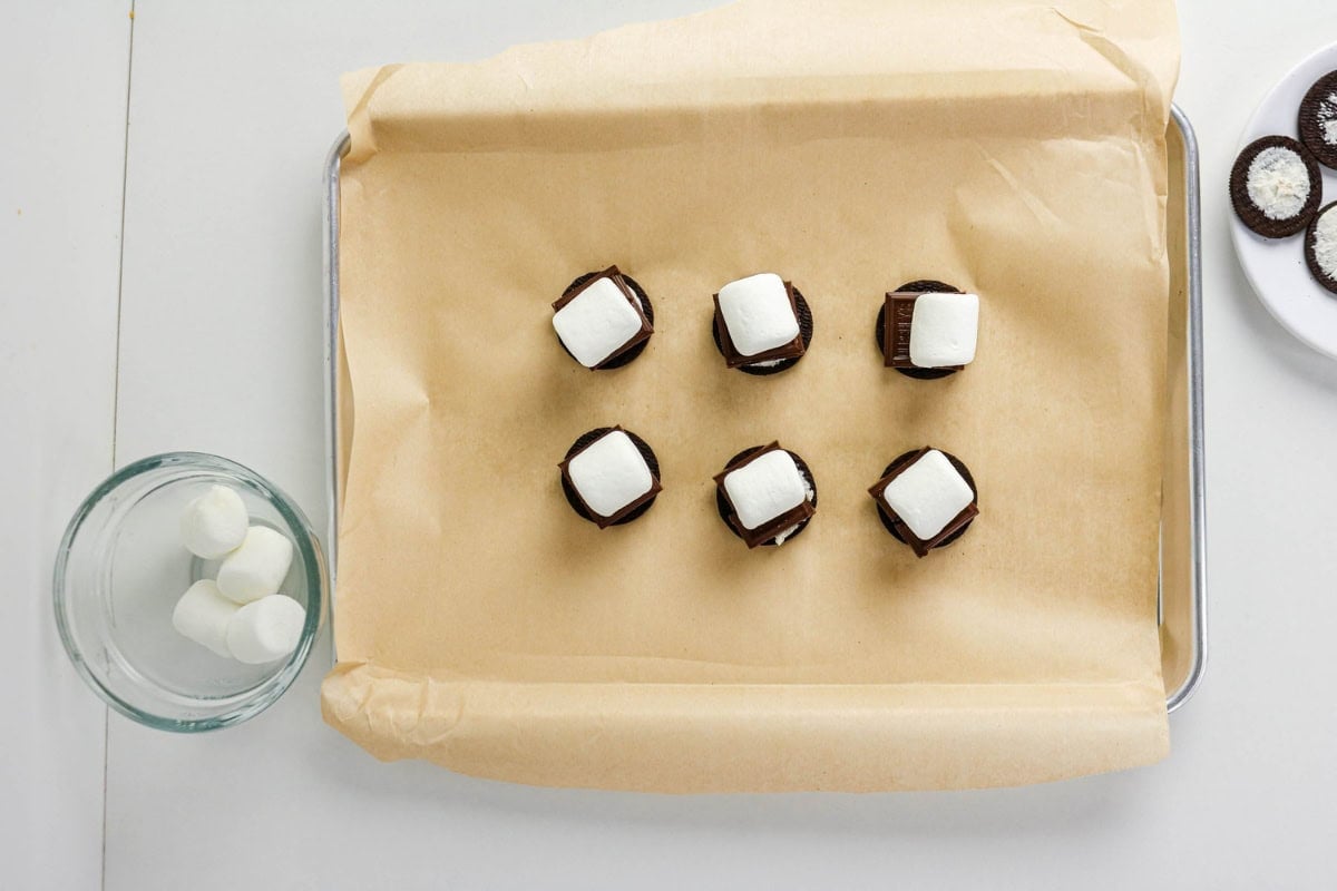 Oreo cookies topped with chocolate and marshmallows on a parchment-lined tray.