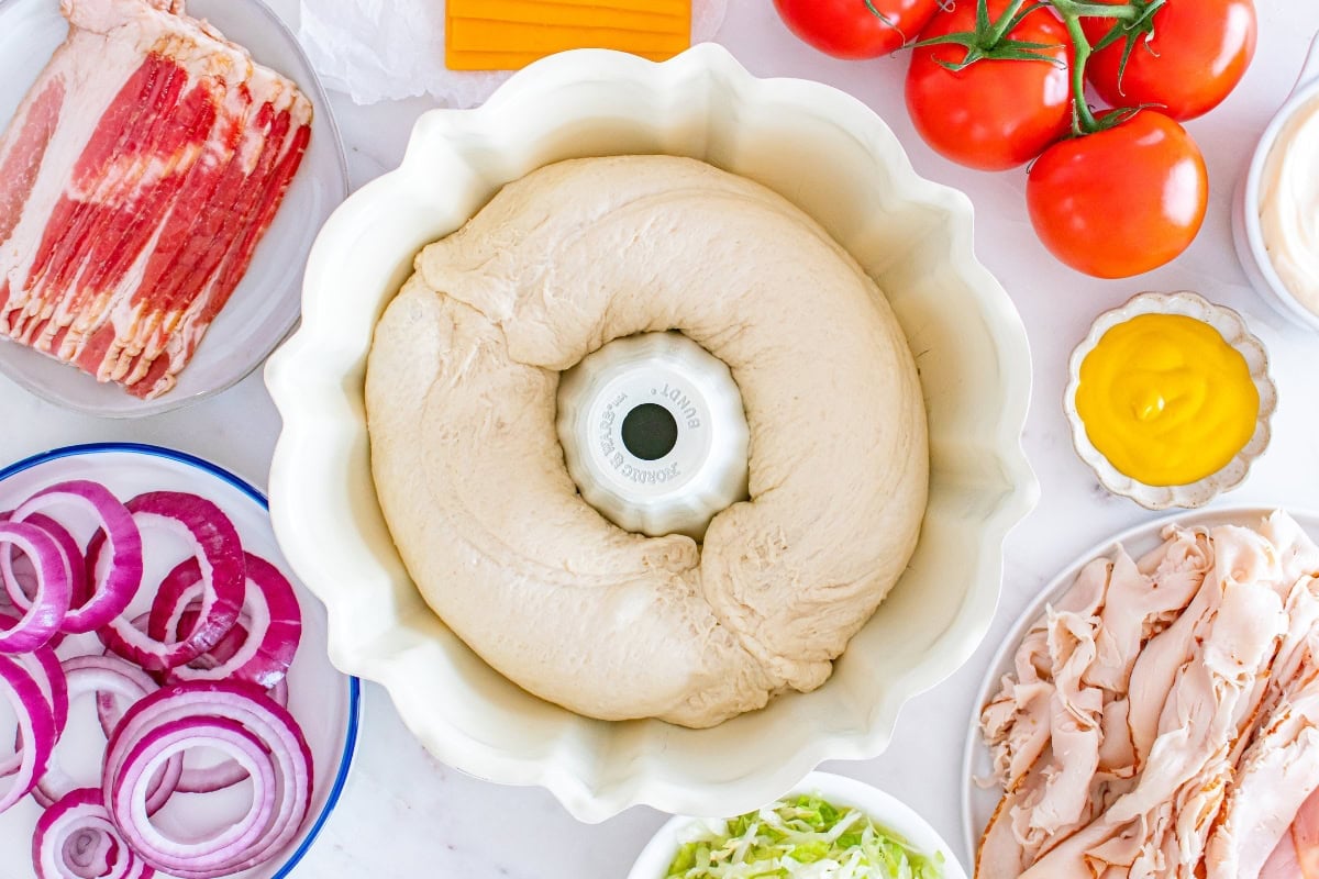 Bundt pan with dough surrounded by bacon, onions, turkey, tomatoes, and mustard.