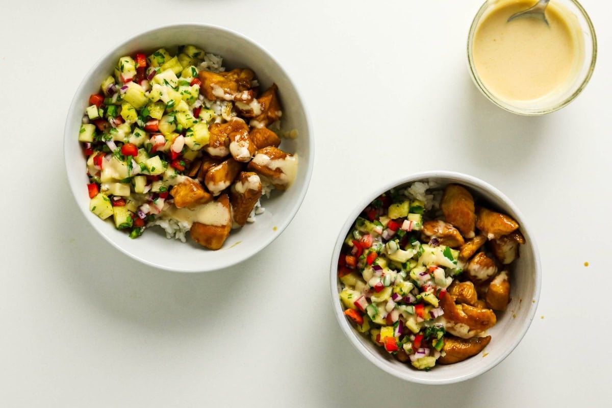 Two bowls of rice with diced chicken and vegetable salsa, plus a cup of sauce.