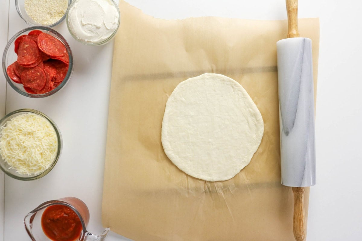 Pizza dough, sauce, cheese, pepperoni, and rolling pin on parchment, ready to make.
