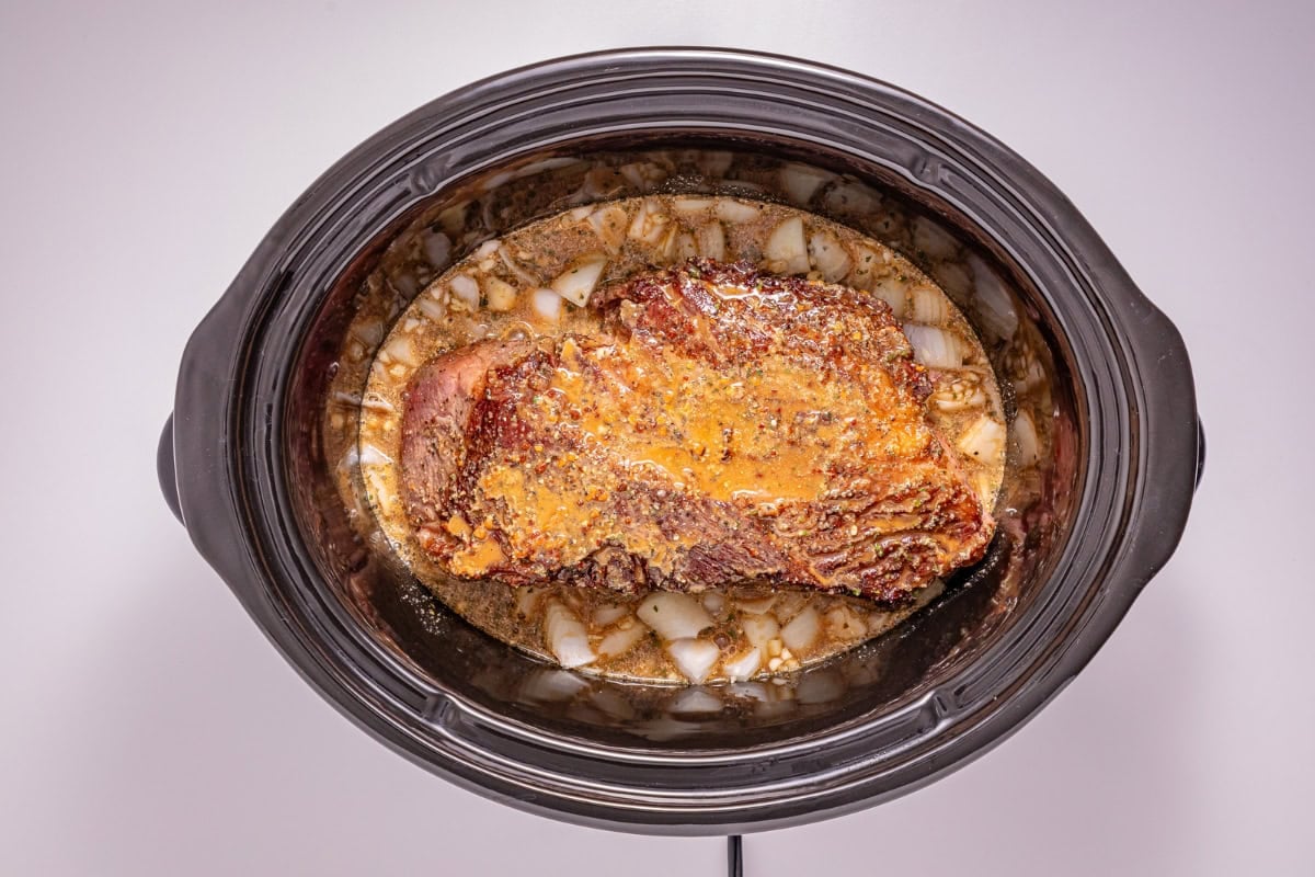 Seasoned meat with onions in a slow cooker filled with broth, top-down view.
