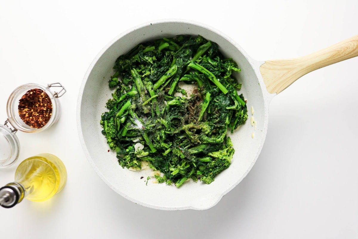 Broccoli rabe cooking in a white skillet with oil and red pepper flakes nearby.