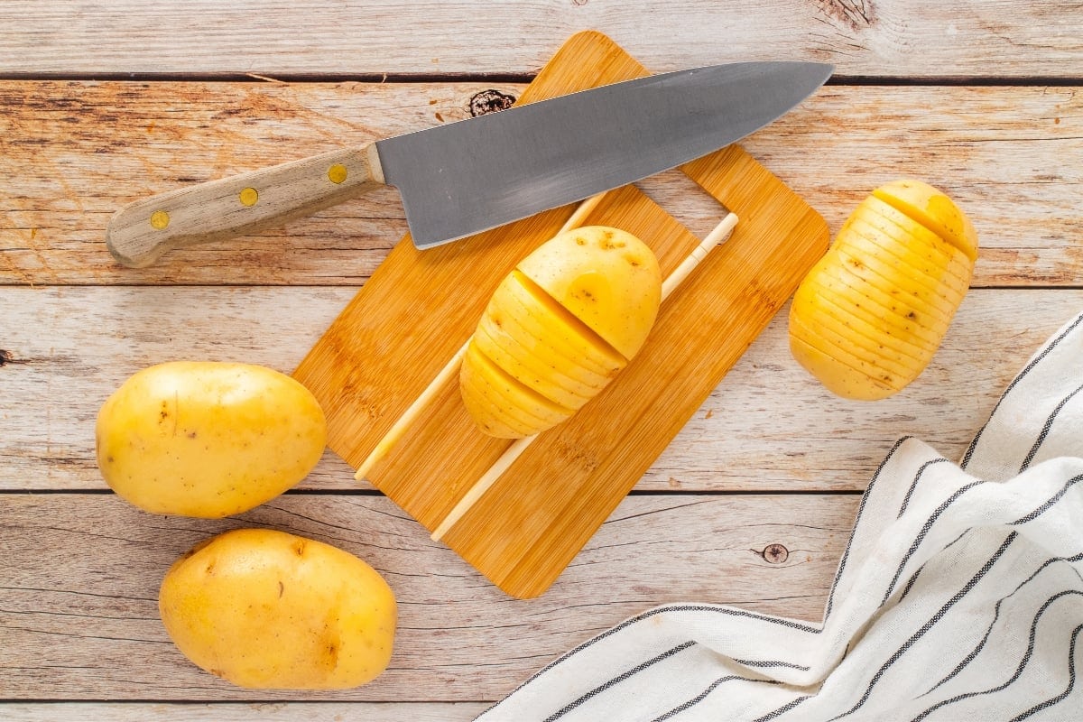Sliced potato on a cutting board with a knife and whole potatoes nearby.