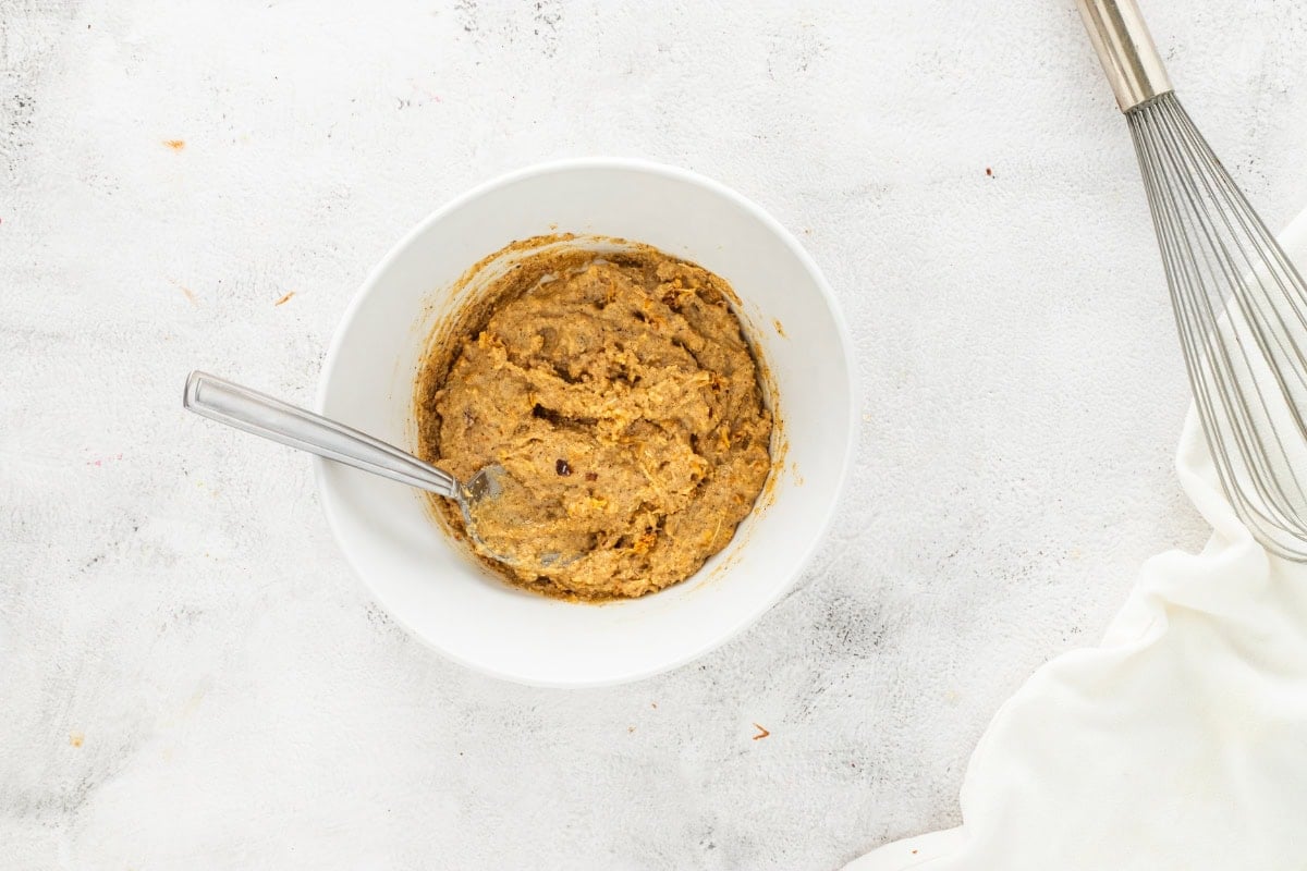 Bowl of cookie dough with a spoon, beside a whisk and white cloth.