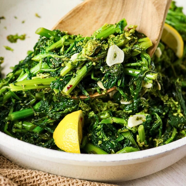 Cooked broccoli rabe with garlic slices and lemon wedges in a white dish.