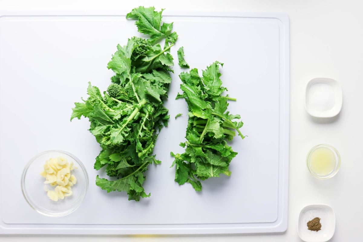 Chopped greens on a board with garlic, salt, pepper, and lemon juice in bowls.