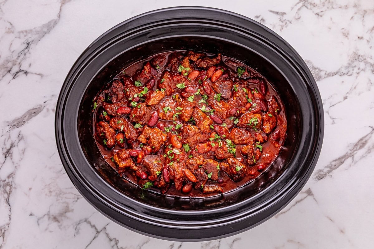 Slow cooker filled with chili topped with chopped herbs on a marble surface.