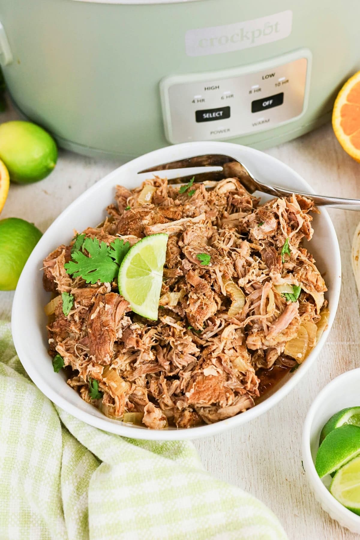 Bowl of shredded pork garnished with lime and cilantro near a slow cooker.