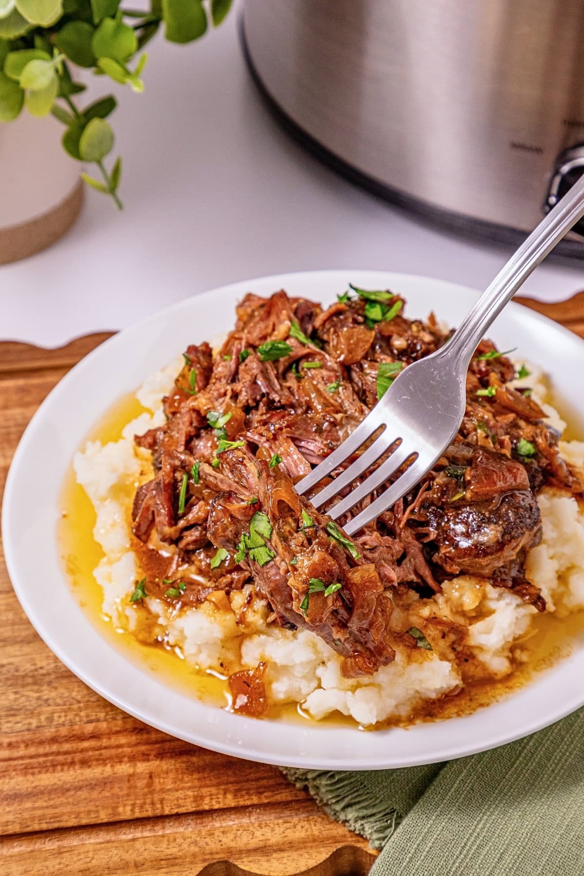 Fork shredding pot roast over mashed potatoes with gravy on a white plate.