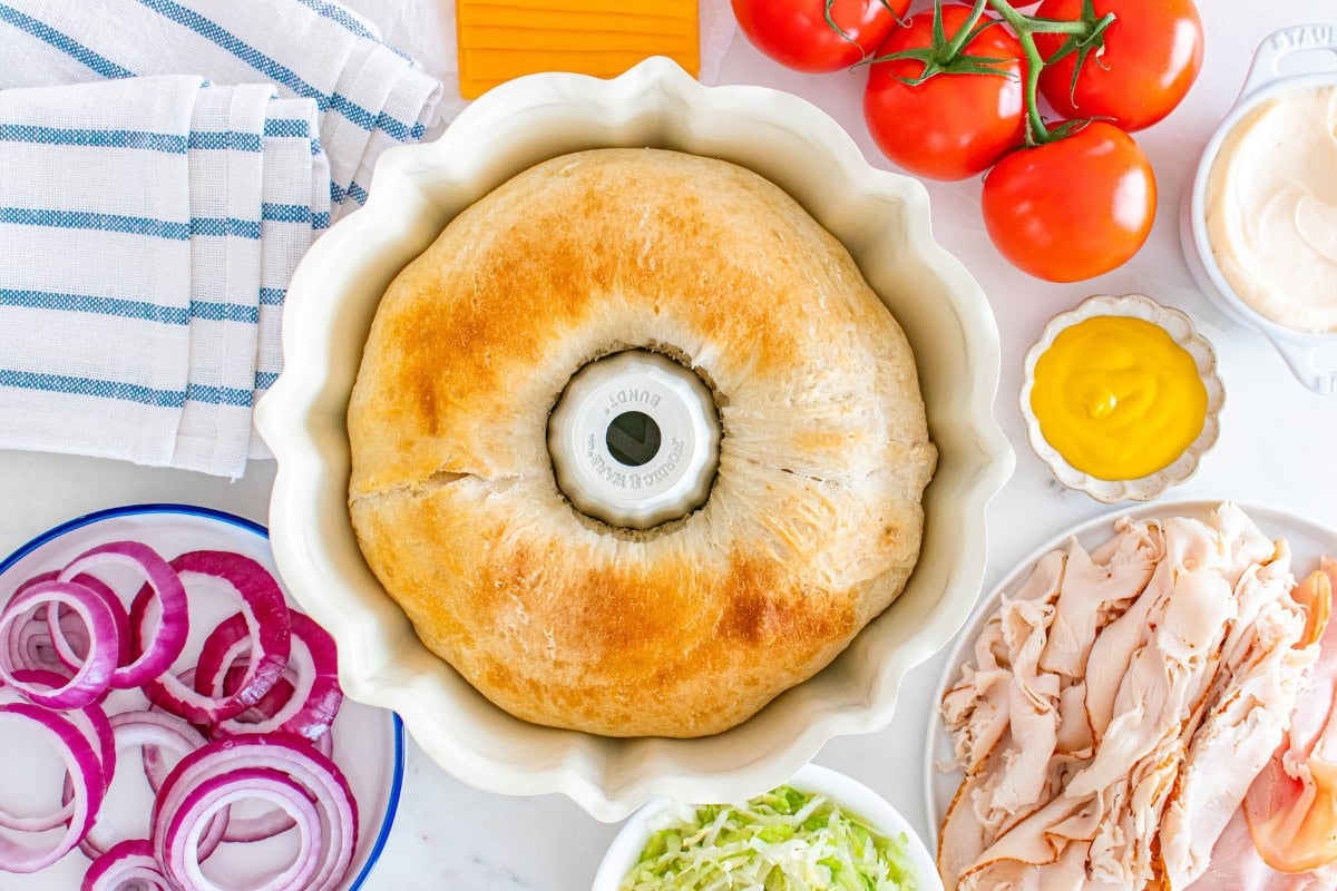 Round baked bread in a bundt pan, surrounded by sandwich fixings and veggies.