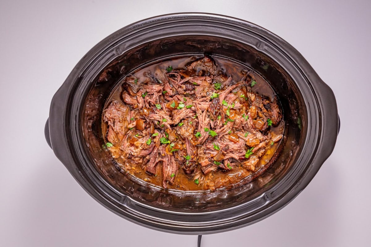 Shredded beef with sauce and herbs in a black slow cooker on a white table.