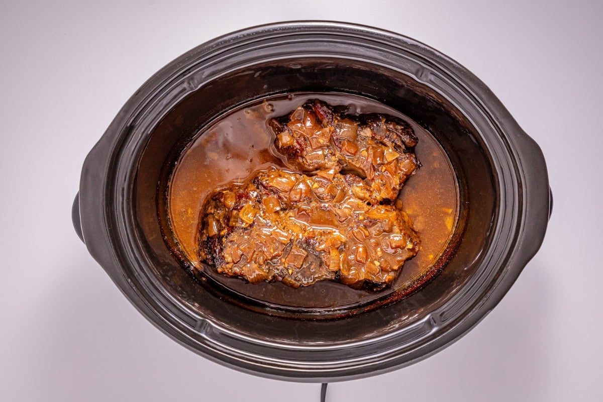 Slow-cooked meat in sauce inside a black crockpot, viewed from above.