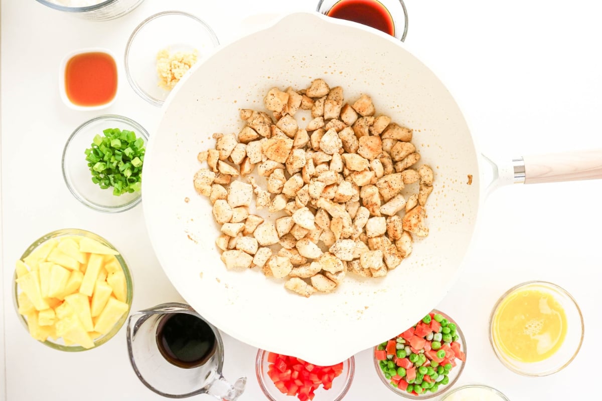 Diced chicken cooking in a white pan surrounded by bowls of chopped ingredients.