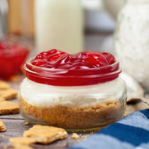 Jar of cherry cheesecake with graham cracker crust on a table.