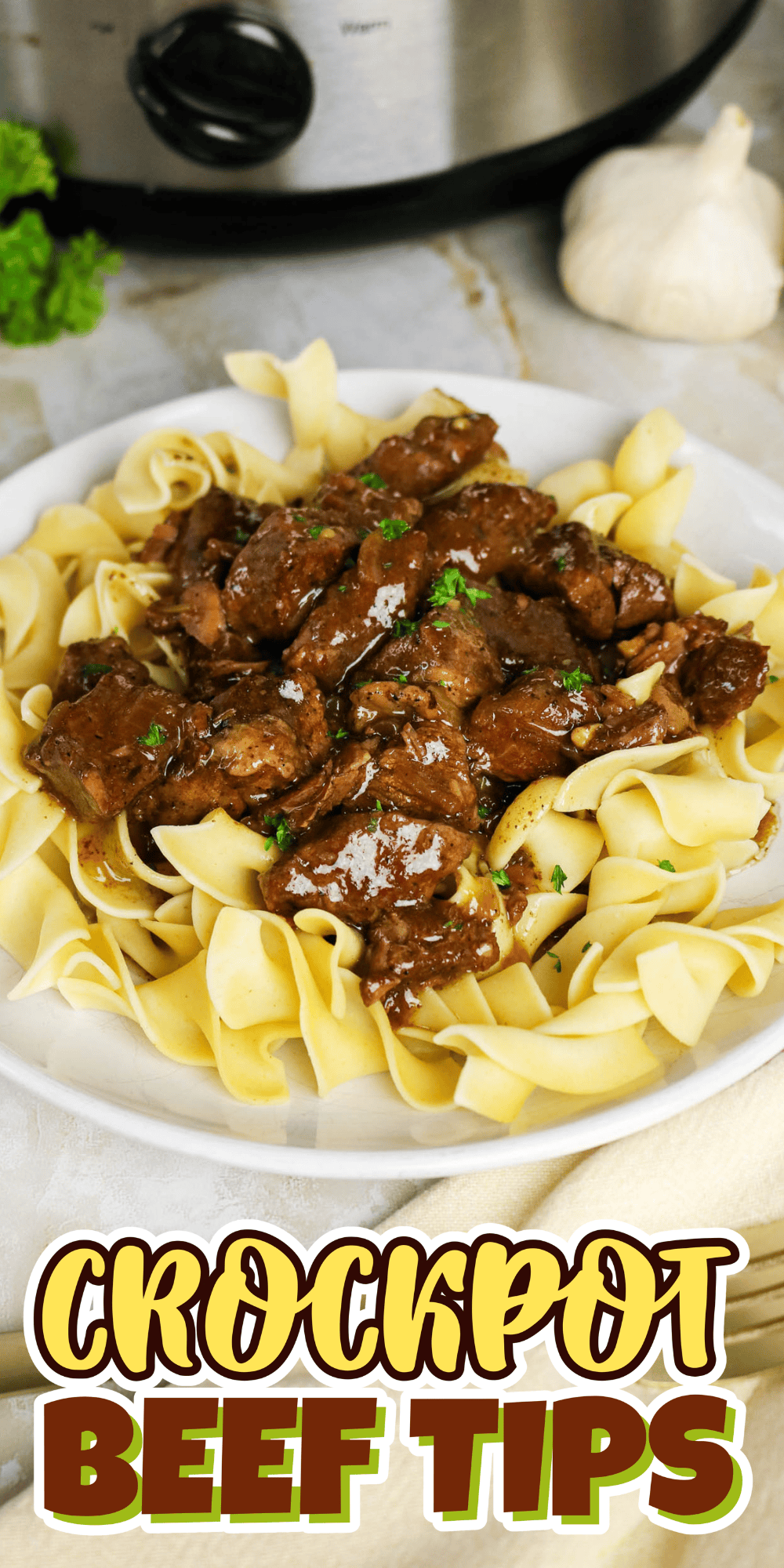 Beef tips in gravy over egg noodles on a white plate, slow cooker in background.