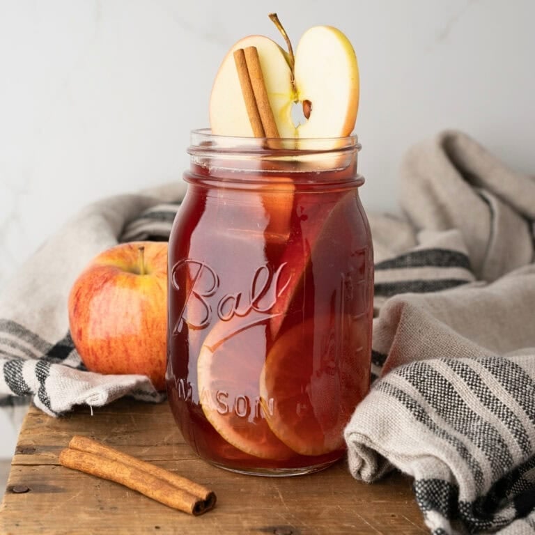 Mason jar of apple cider with apple slices and cinnamon sticks on a table.