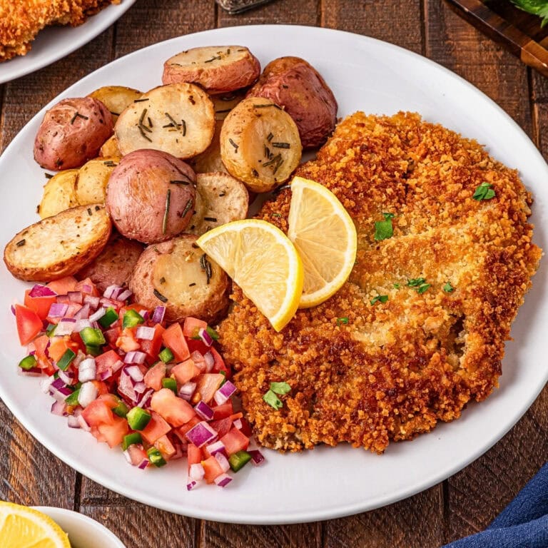 Breaded cutlet, roasted potatoes, lemon slices, and diced veggie salad on plate.