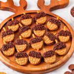 Acorn-shaped cookies with chocolate and sprinkles on a round wooden tray.