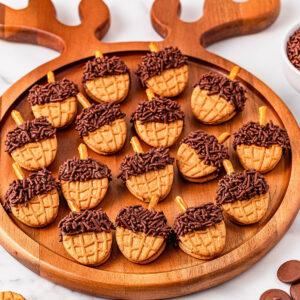 Acorn-shaped cookies with chocolate and sprinkles on a round wooden tray.