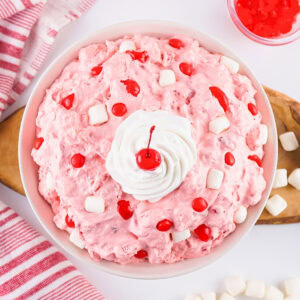 Pink dessert with whipped cream, cherries, and marshmallows in a white bowl.