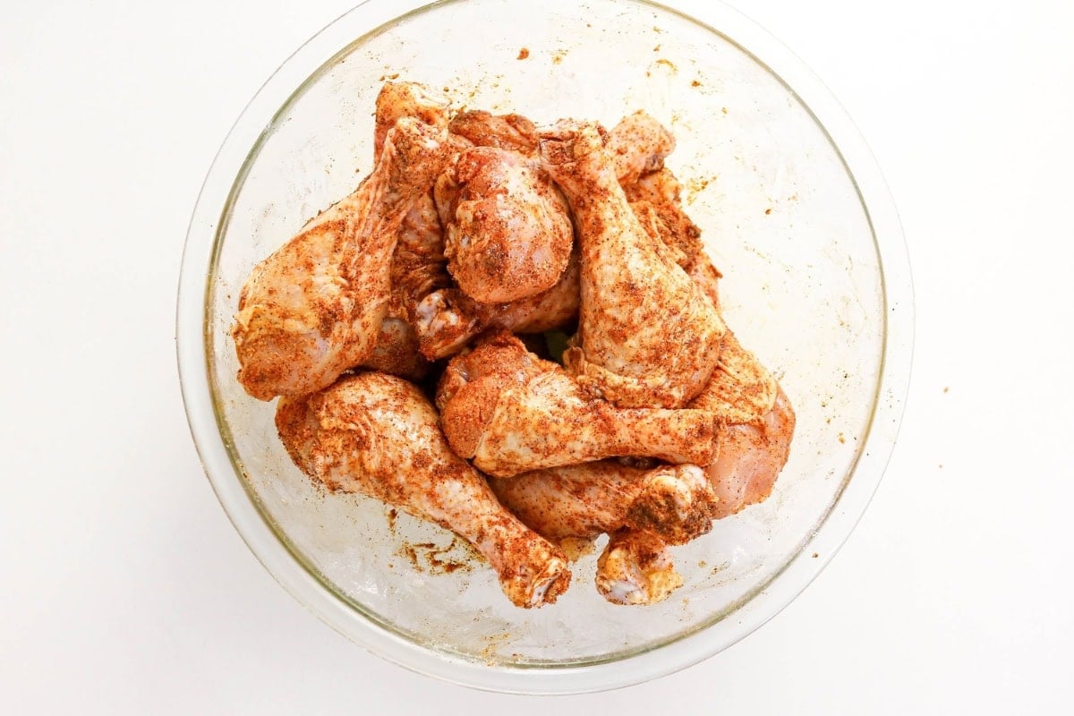 Raw chicken drumsticks coated with spices in a clear glass bowl.