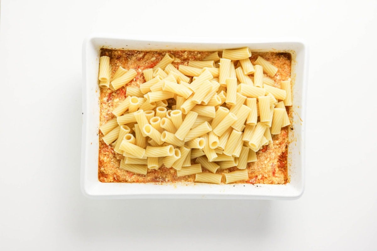Unmixed rigatoni pasta and sauce in a white baking dish on a white background.