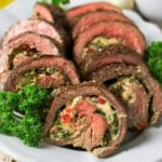 Sliced stuffed steak rolls with cheese, herbs, and vegetables on a white plate.