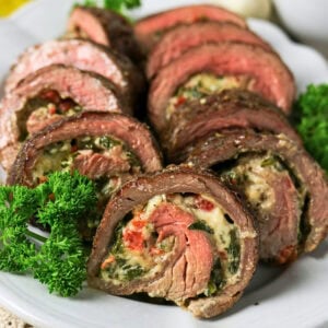 Sliced stuffed steak rolls with cheese, herbs, and vegetables on a white plate.