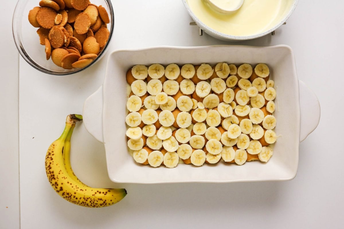 Banana slices in a dish with a banana, cookies, and a bowl of pudding nearby.