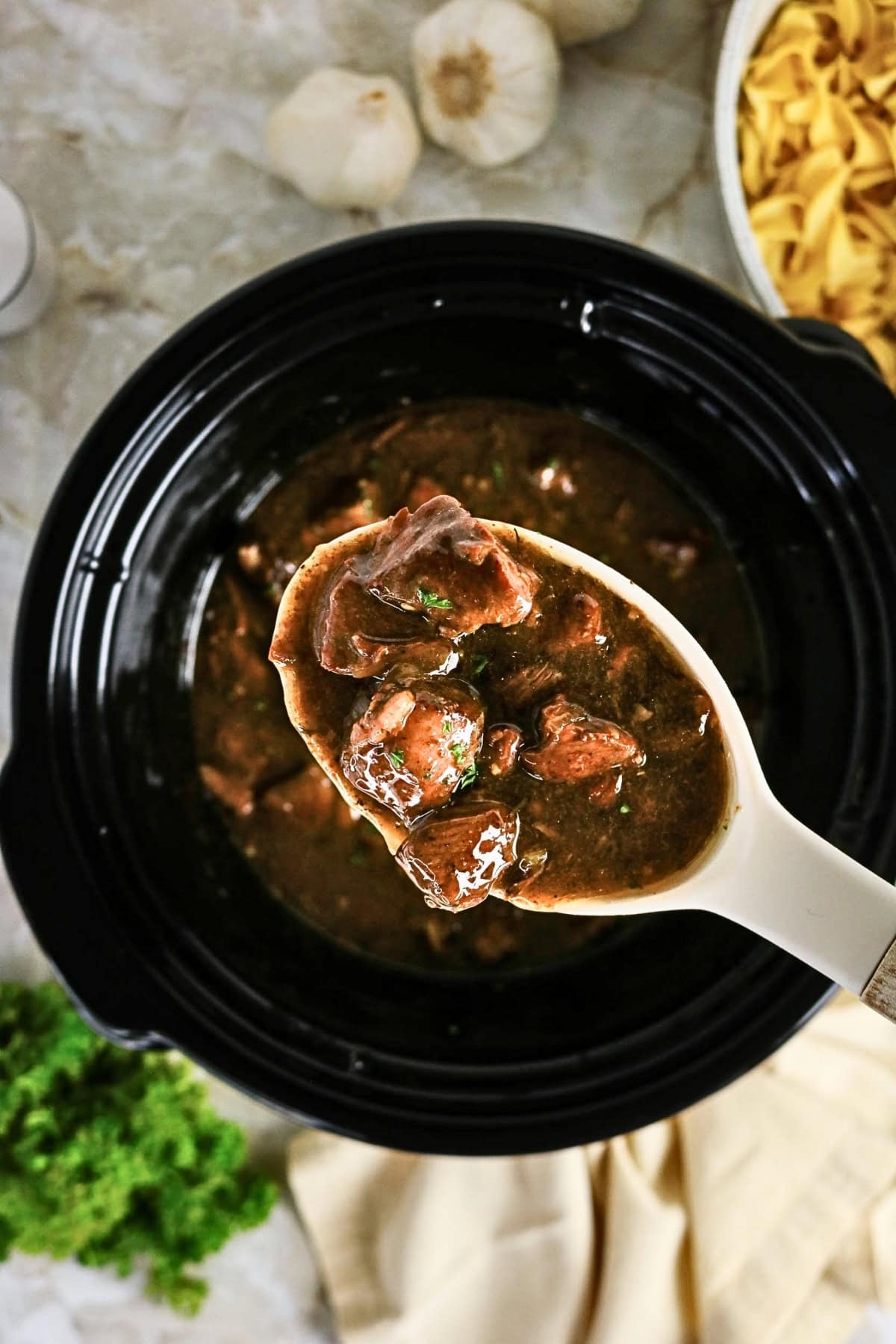 Ladle of beef stew over a slow cooker, with herbs and noodles nearby.