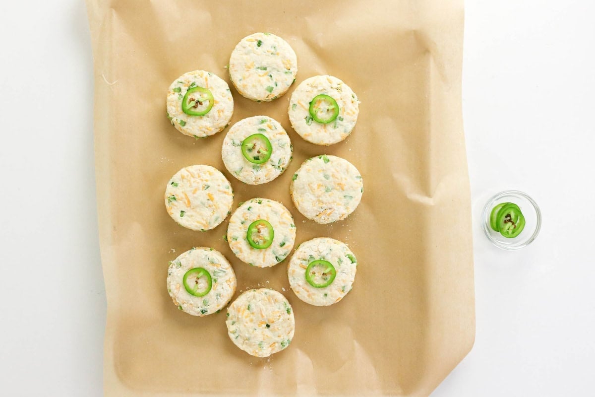 10 uncooked jalapeño biscuits on parchment, with a small jar of jalapeño slices.