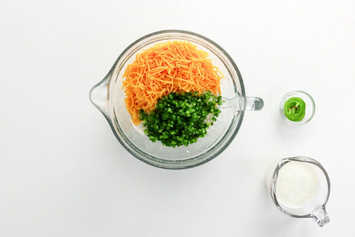 Shredded cheese, chopped jalapeños in a bowl, with milk and jalapeños on side.
