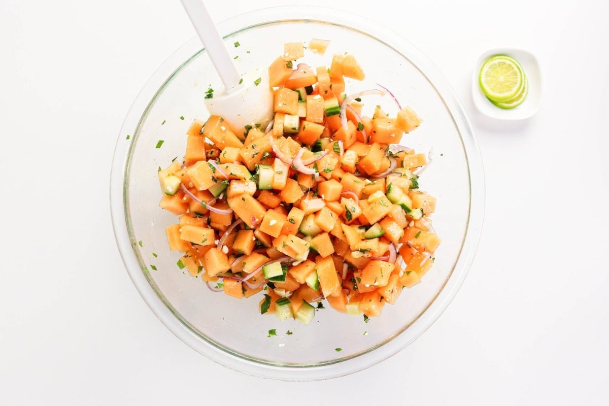 A bowl of diced cantaloupe salad with cucumber and herbs, plus lime slices.