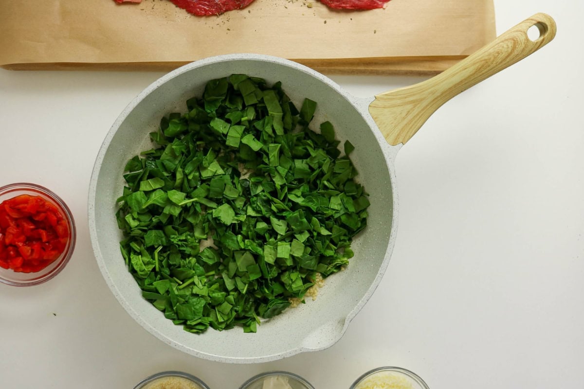 Chopped spinach cooking in a white pan, with diced red pepper on the side.