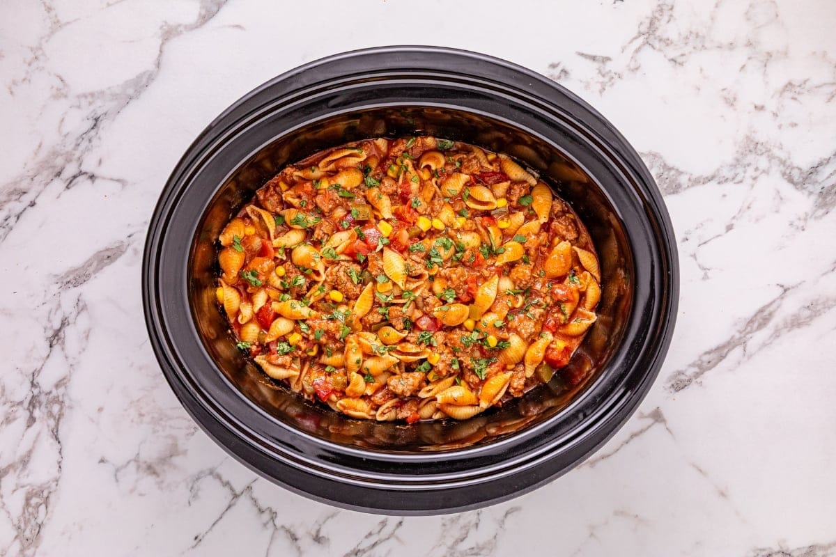 Shell pasta with meat sauce and herbs in a black slow cooker on marble surface.