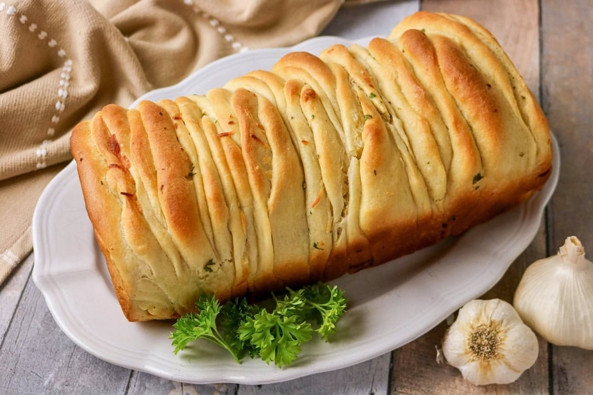 Stacked garlic bread loaf on a white platter, parsley and garlic nearby.